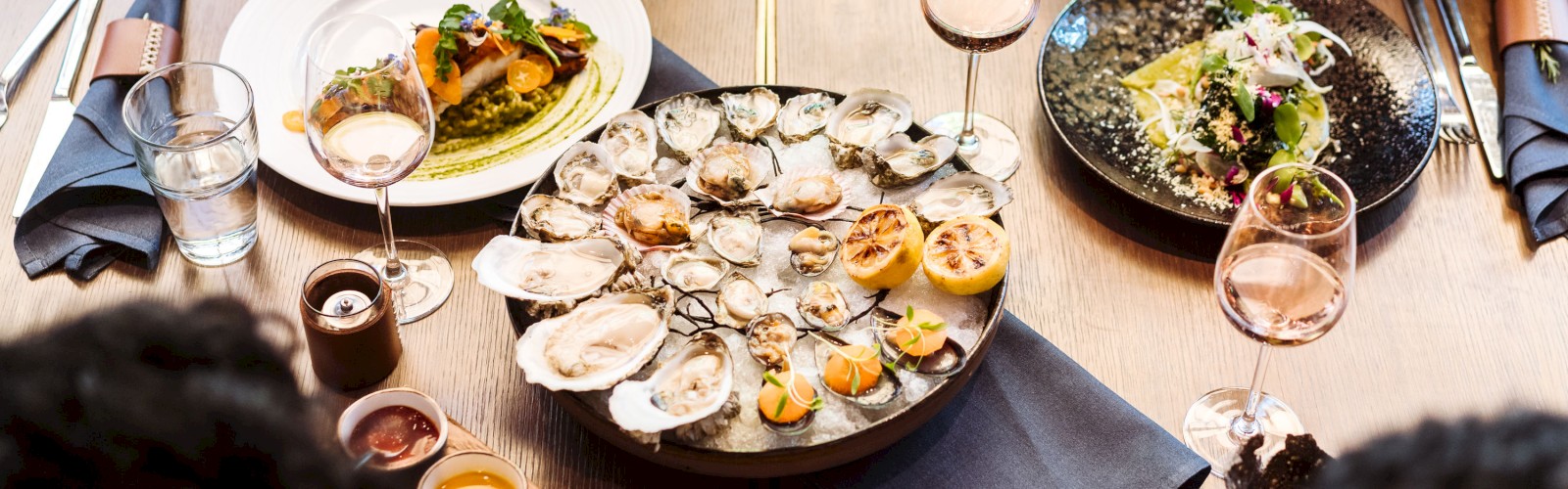 A table set for a meal with oysters, a fish dish, a salad, sauces, wine glasses, and napkins, with people seated around it.