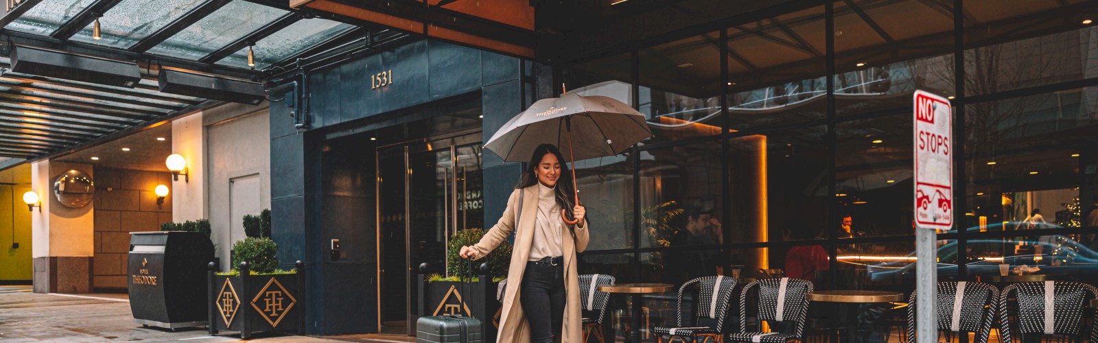 A woman holding an umbrella is walking with rolling luggage outside Hotel Theodore on a rainy day. She is dressed in a long coat and casual attire.