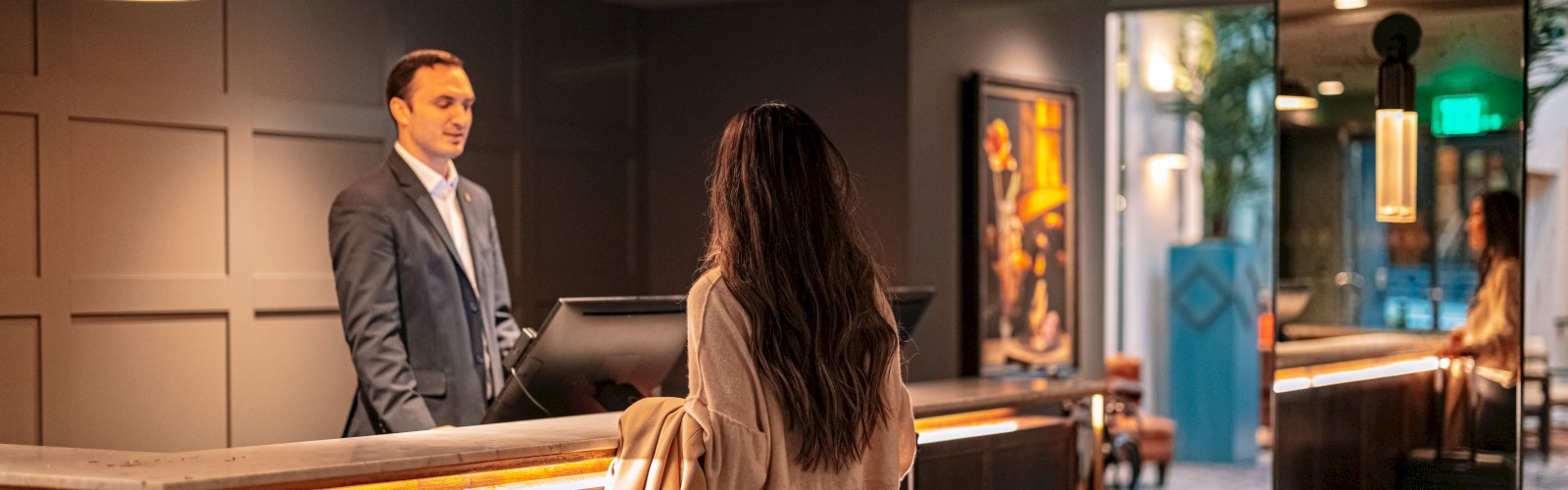 A woman standing at a hotel reception desk interacts with the receptionist; she has luggage beside her and a coat in hand, completing the check-in.