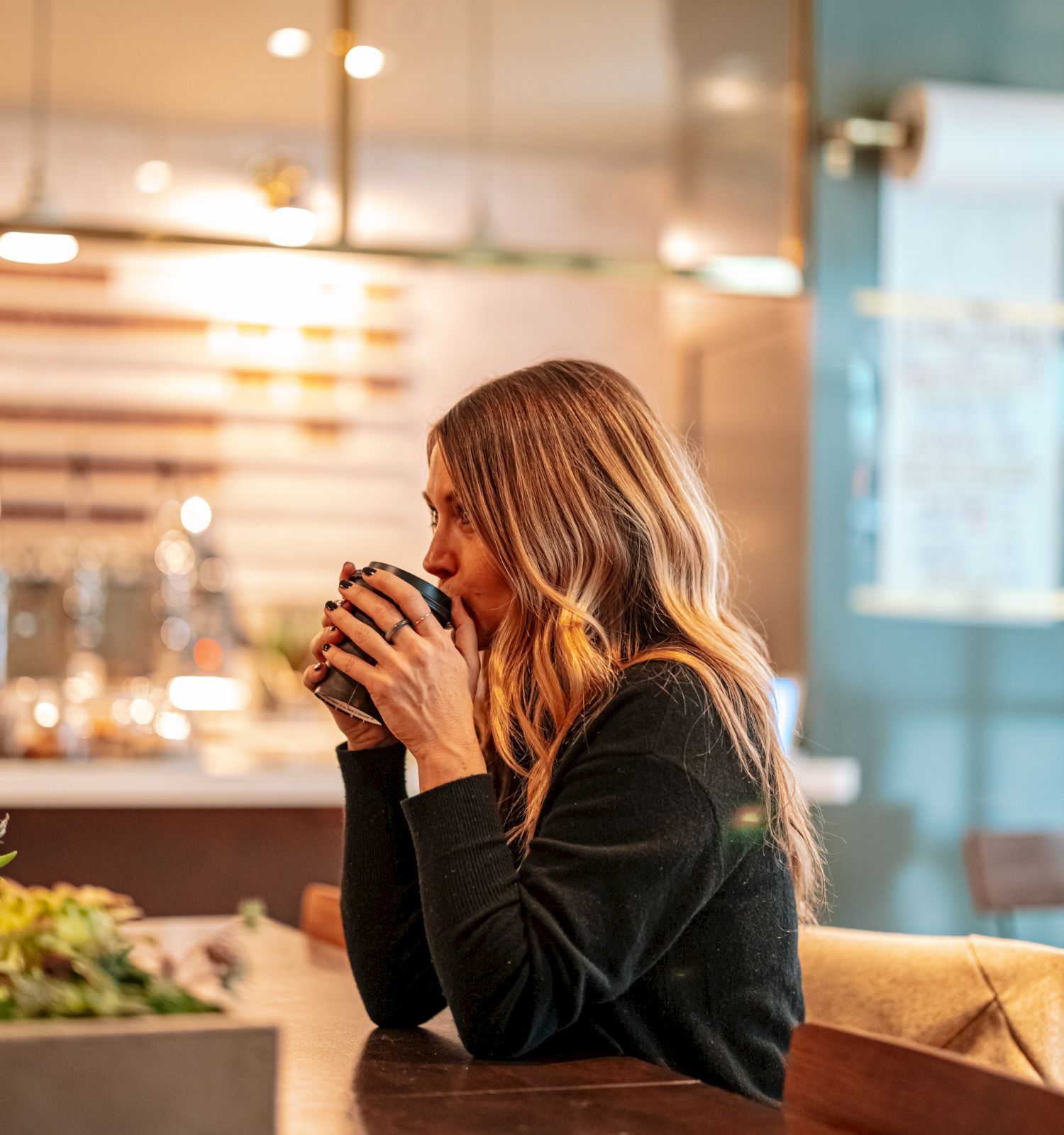 A woman with long hair sits at a wooden table, holding a cup and gazing thoughtfully. The background features indoor lighting and decorations.