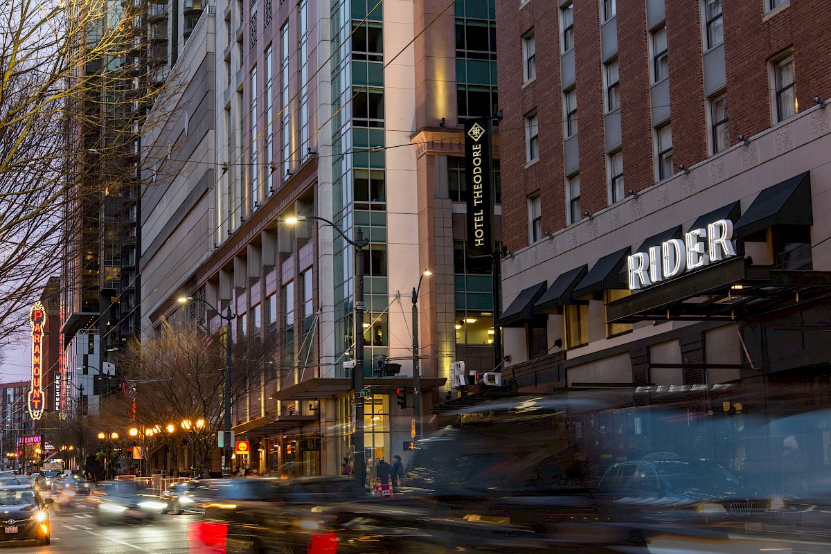 A busy urban street with cars, high-rise buildings, Hotel Theodore, Rider sign, and streetlights glowing at dusk.