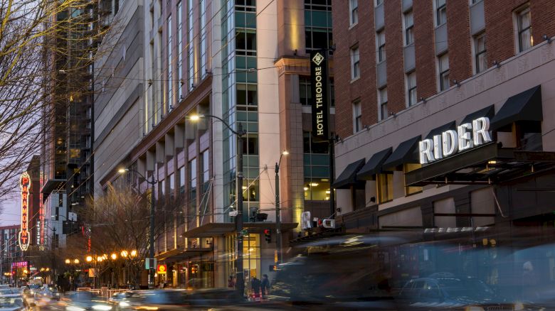 A busy urban street with cars, high-rise buildings, Hotel Theodore, Rider sign, and streetlights glowing at dusk.