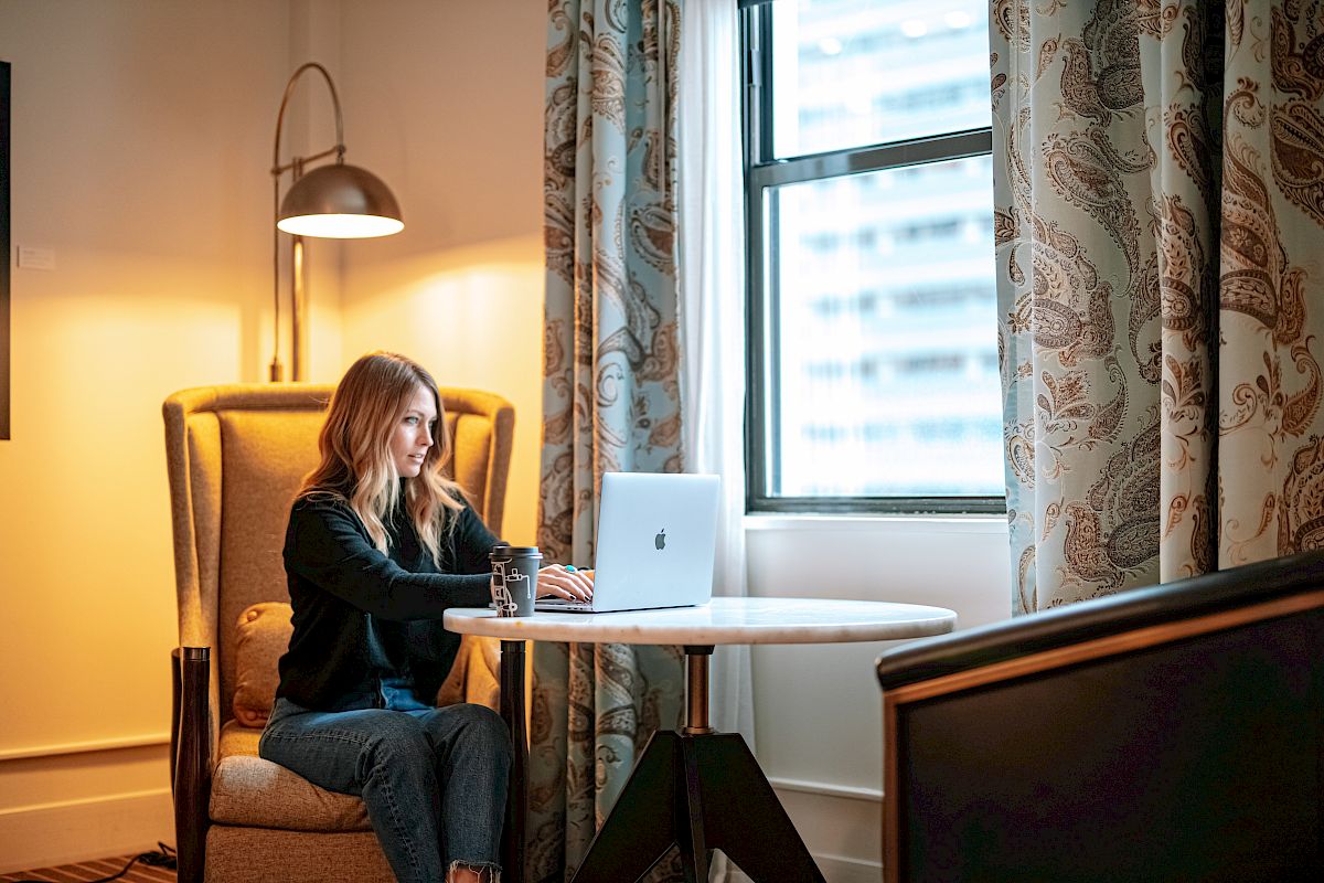 A woman is seated in a room with a laptop on a small round table, a glass of water beside her, near a window with patterned curtains.