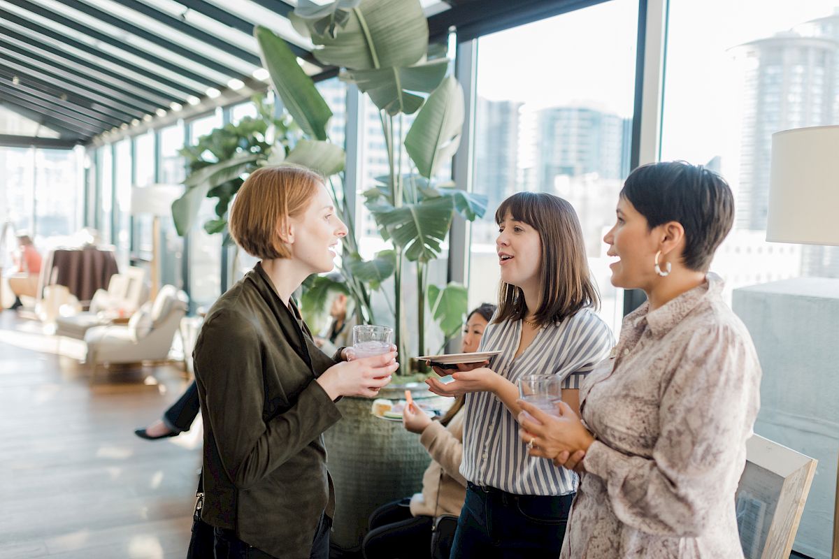 Three women are standing in a bright room with large windows and plants, chatting and holding drinks. A few people are seated in the background.