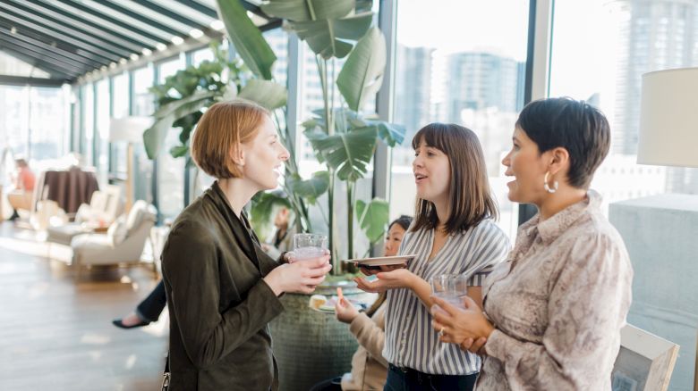 Three women are standing in a bright room with large windows and plants, chatting and holding drinks. A few people are seated in the background.