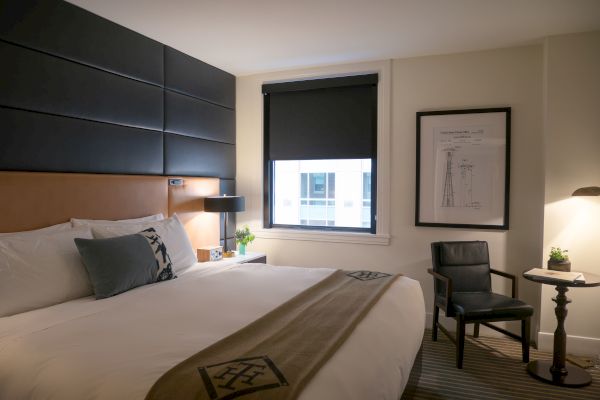 A neatly arranged hotel room with a bed, side table, chair, window, lamp, and framed picture on the wall ends the sentence.