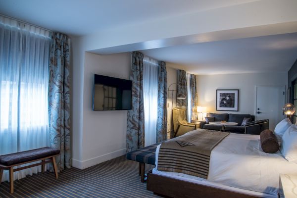 The image shows a modern hotel room with a large bed, a seating area, a wall-mounted TV, and large windows with curtains.