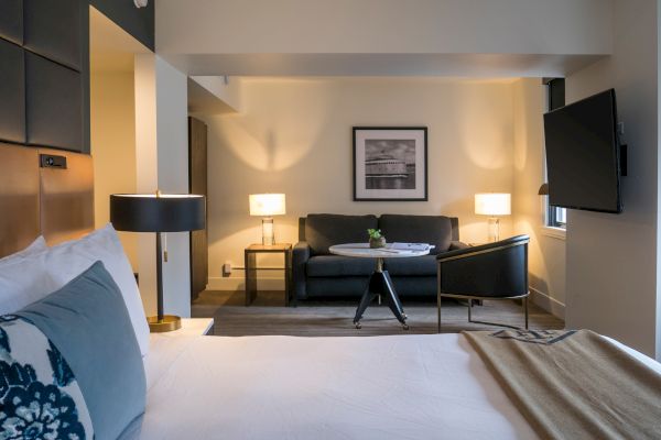 A modern hotel room with a bed, side table, and lamp in the foreground. A seating area with a couch, table, and TV in the background.