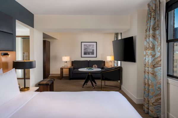 A modern hotel room with a bed, a seating area with a sofa, a TV, lamps, wall art, and large windows with curtains, bright and inviting.