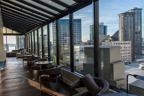 This image shows a glass-enclosed lounge area with modern furniture and a view of city buildings through large windows, under a clear blue sky.