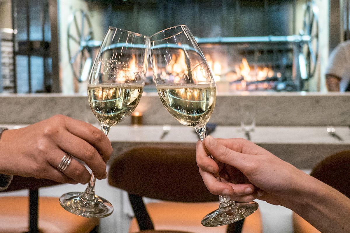 Two people clink glasses of white wine in a cozy restaurant setting with a fireplace in the background.