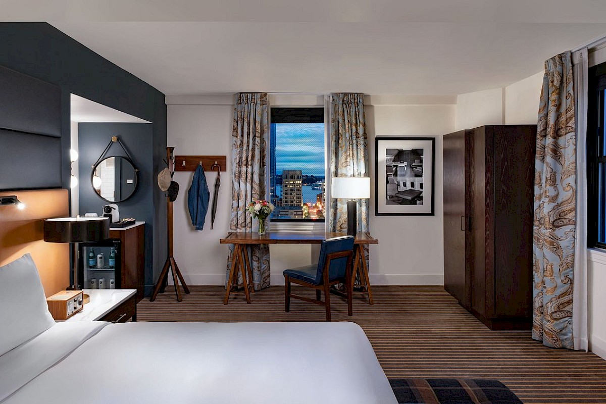 The image shows a stylish hotel room with a bed, desk, chair, wardrobe, and window view of city lights, decorated in modern tones.