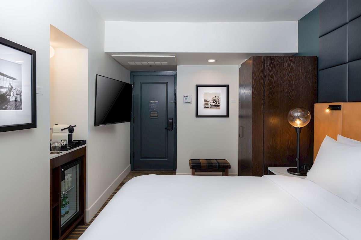 This image shows a modern hotel room with a large bed, wall-mounted TV, mini fridge, and stylish decor, including framed photos and a globe-style lamp.