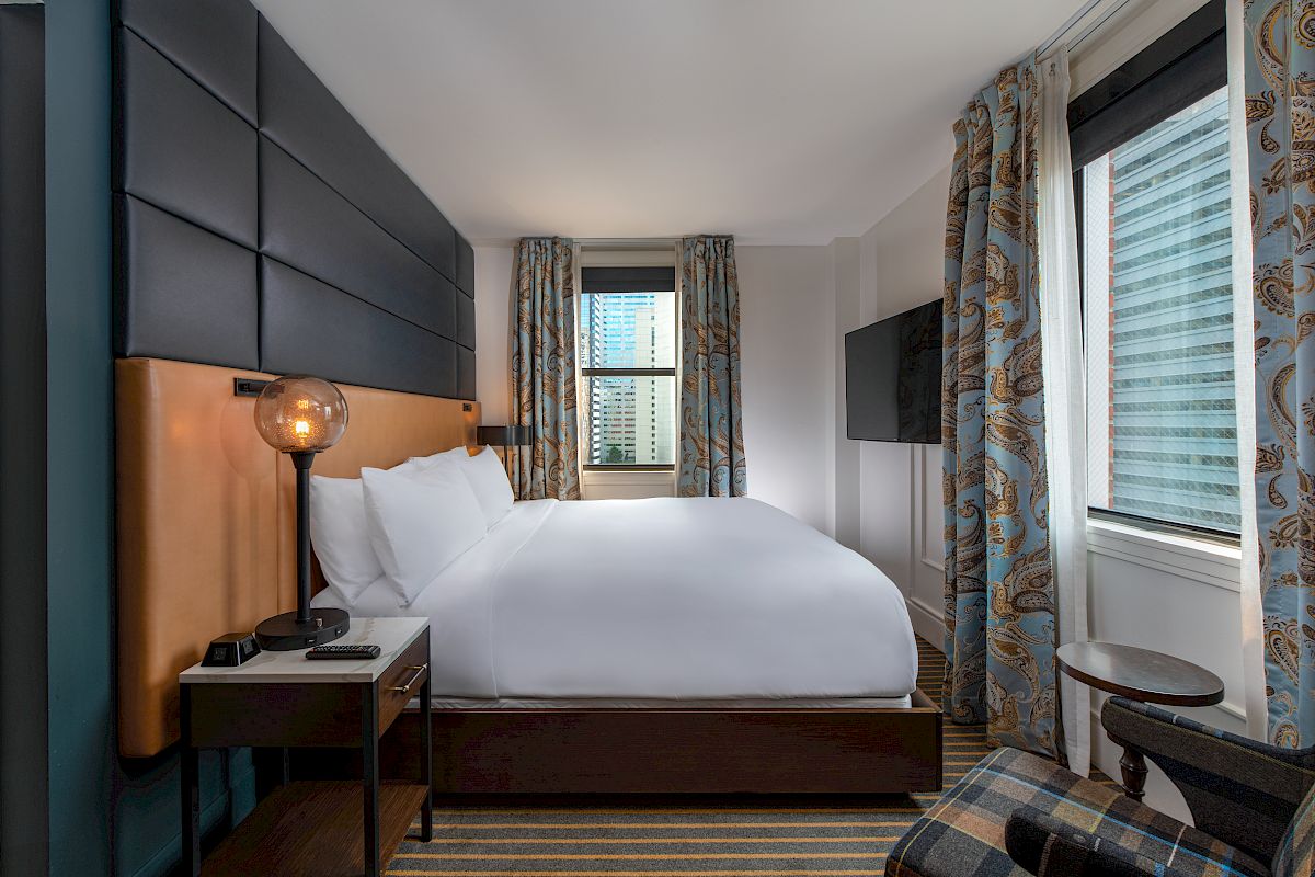The image shows a modern hotel room with a neatly made bed, bedside table, lamp, wall-mounted TV, and a chair by the window with patterned curtains.