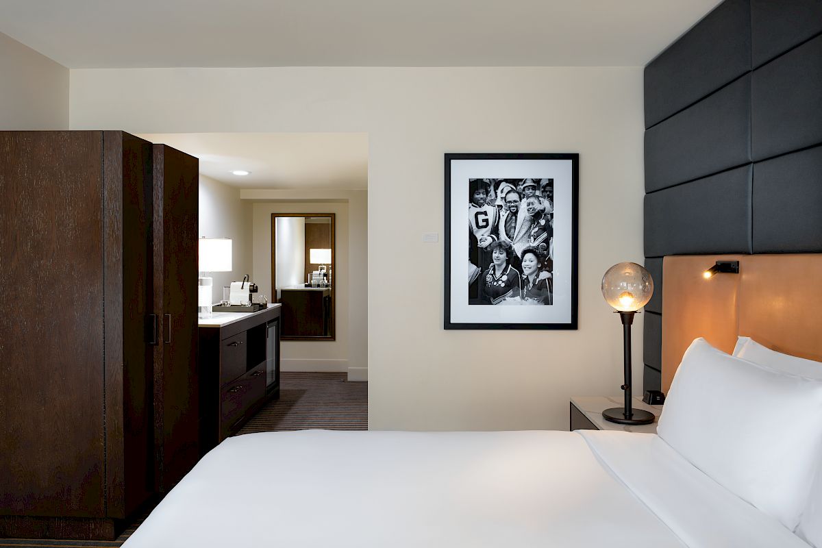 A neat hotel room features a white bed, a framed photo on the wall, a wardrobe, and a lit lamp. The room extends into a well-lit area with a desk.