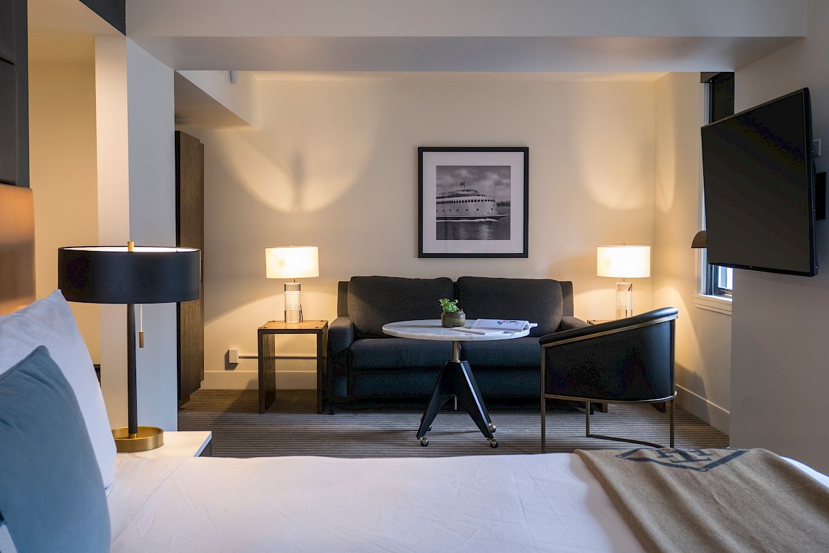 This is a photo of a modern hotel room with a cozy seating area, including a sofa, chairs, coffee table, lamps, and wall art.