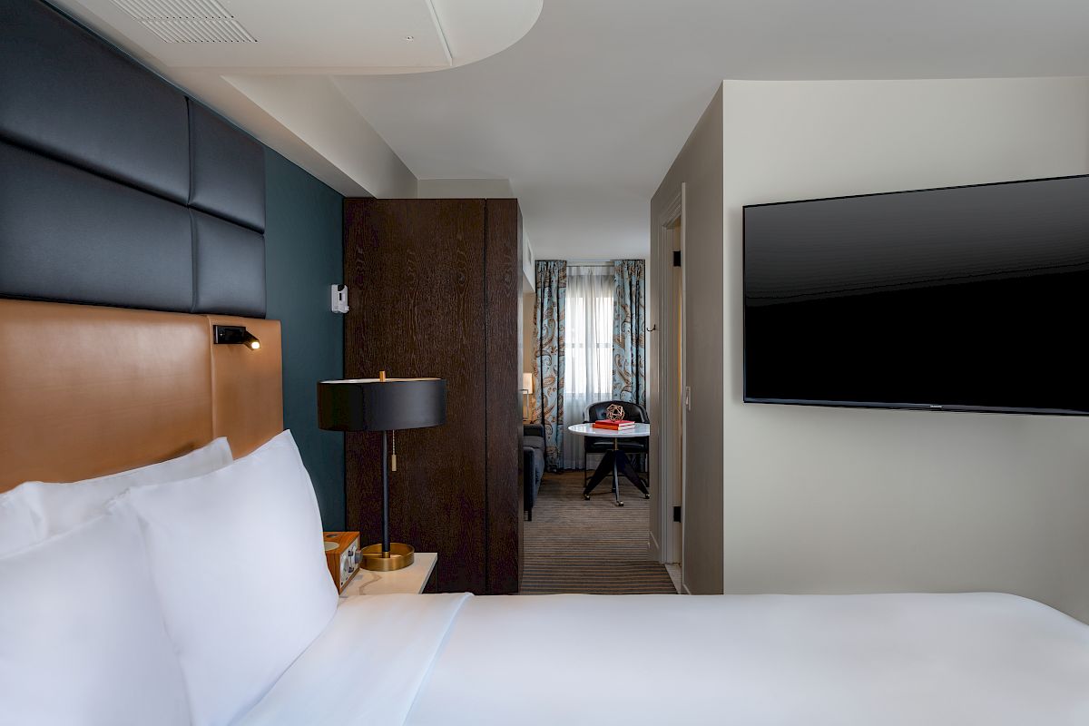 A modern hotel room with a bed, wall-mounted TV, bedside lamp, and a glimpse of a sitting area in the background, ending the sentence.