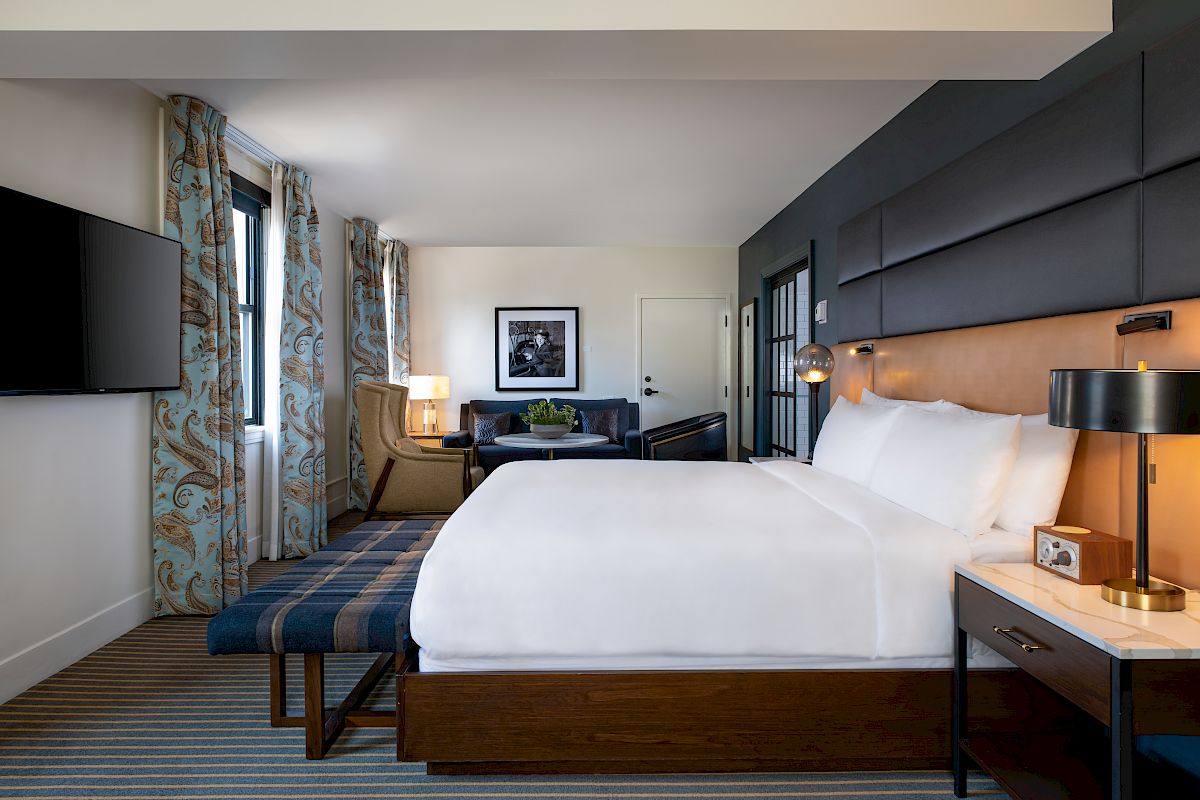 A modern hotel room with a large bed, wall-mounted TV, side tables, sofa, armchairs, and a window with patterned curtains, ending the sentence.