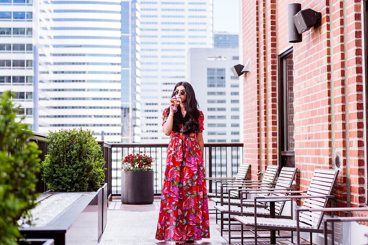 A woman in a red floral dress is standing on a balcony with city skyscrapers in the background, holding a drink, surrounded by outdoor furniture.
