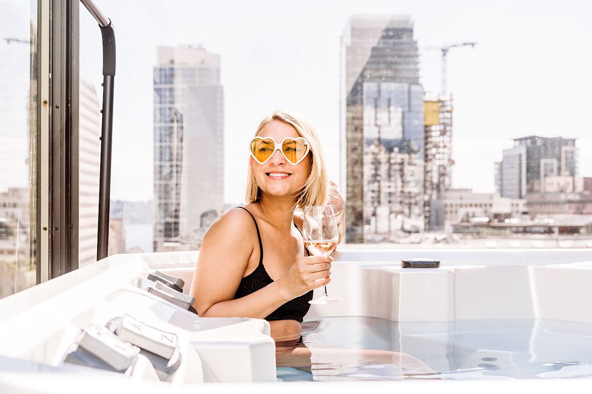 A person in sunglasses is smiling and holding a drink while relaxing in a hot tub on a rooftop with a cityscape in the background.