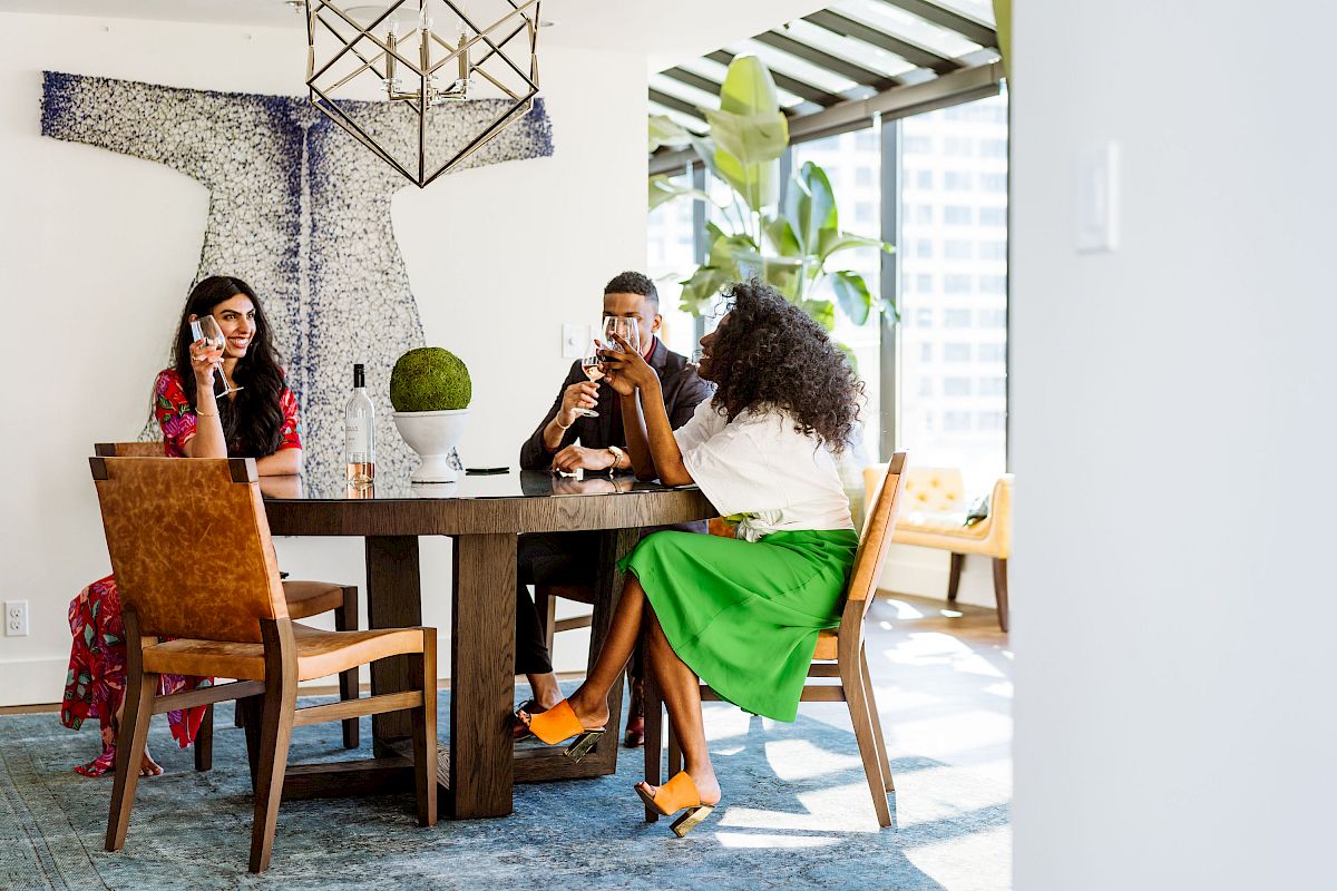 Three people are sitting around a wooden table, drinking and socializing in a bright, modern room with a large window and indoor plants.
