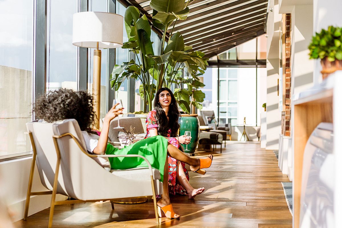Two women are sitting in a sunlit, modern room with large windows, indoor plants, and wooden flooring, enjoying drinks and conversation.