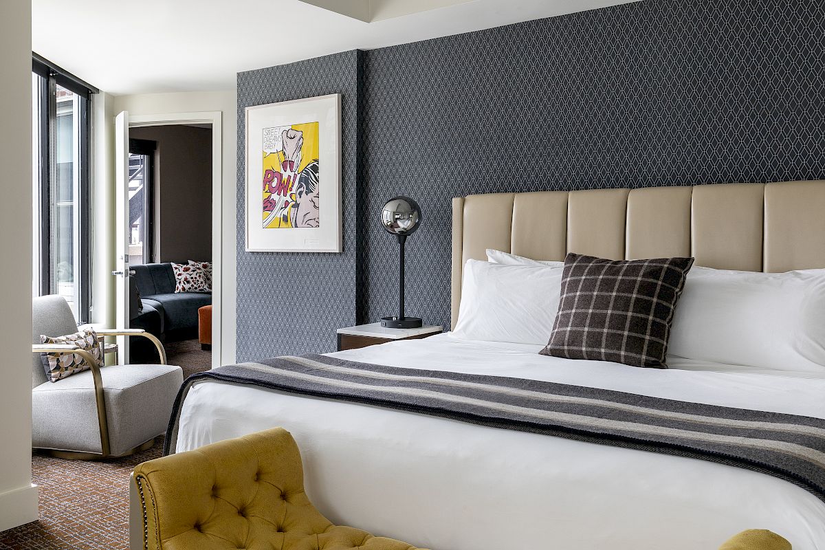 A modern hotel room with a large bed, gray accent wall, artwork, seating area, and floor-to-ceiling windows.