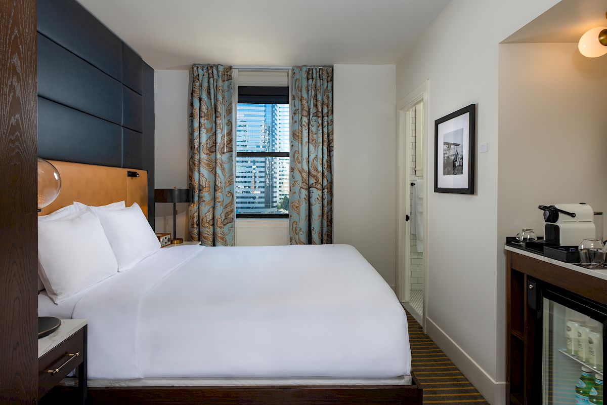 A modern hotel room with a large bed, city view window, patterned curtains, wall art, and a mini fridge and coffee maker in the corner.