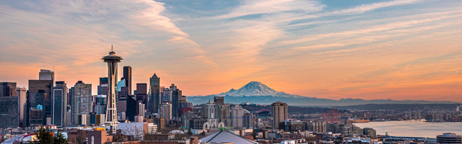 A city skyline with a prominent tower and various buildings, set against a backdrop of a snow-capped mountain under a colorful sky at sunset.