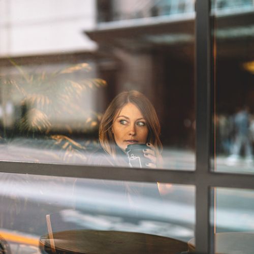 A woman is sitting at a table in a cafe, drinking from a cup and looking out the window with a contemplative expression.