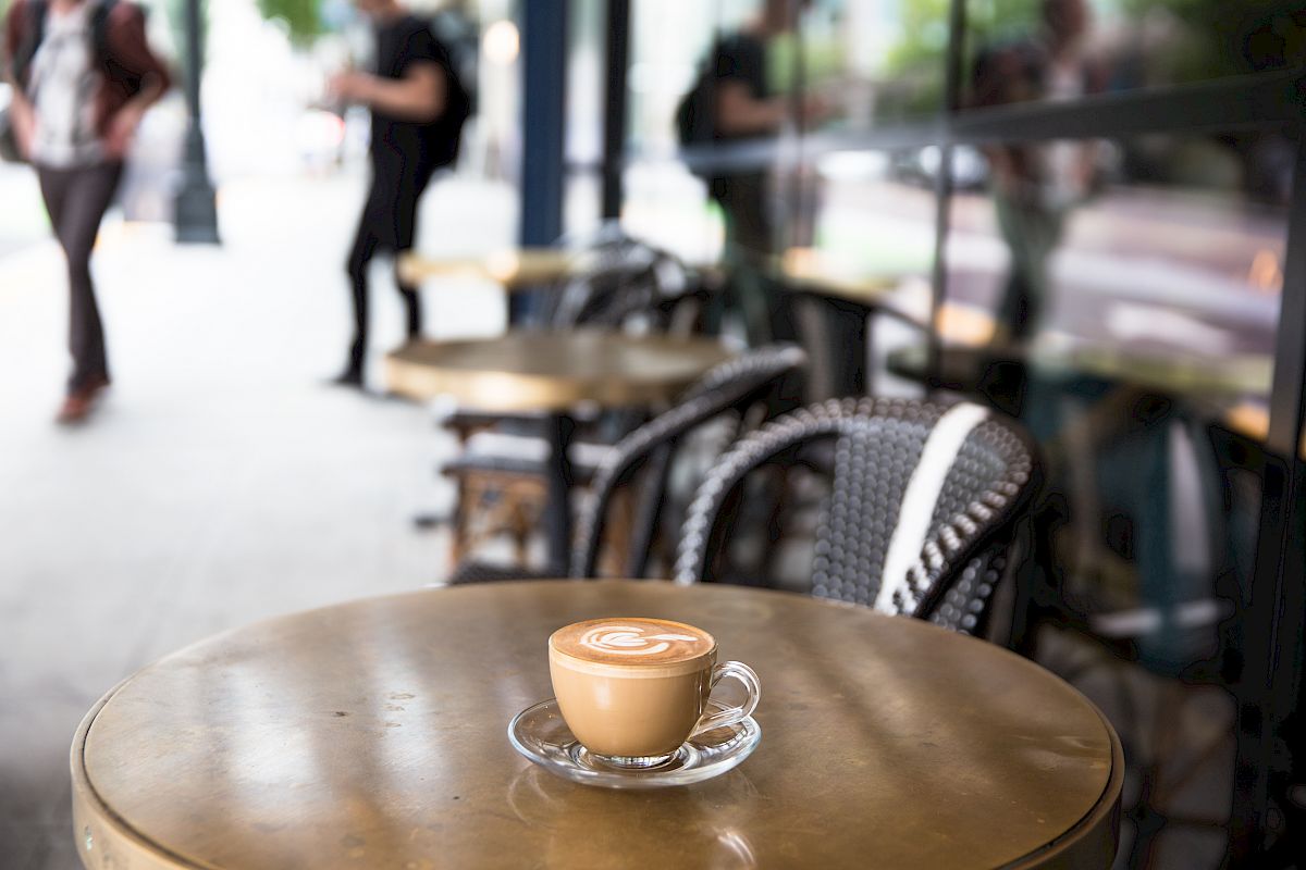 A cup of coffee on an outdoor café table, with empty chairs and blurred pedestrians in the background on a sidewalk.