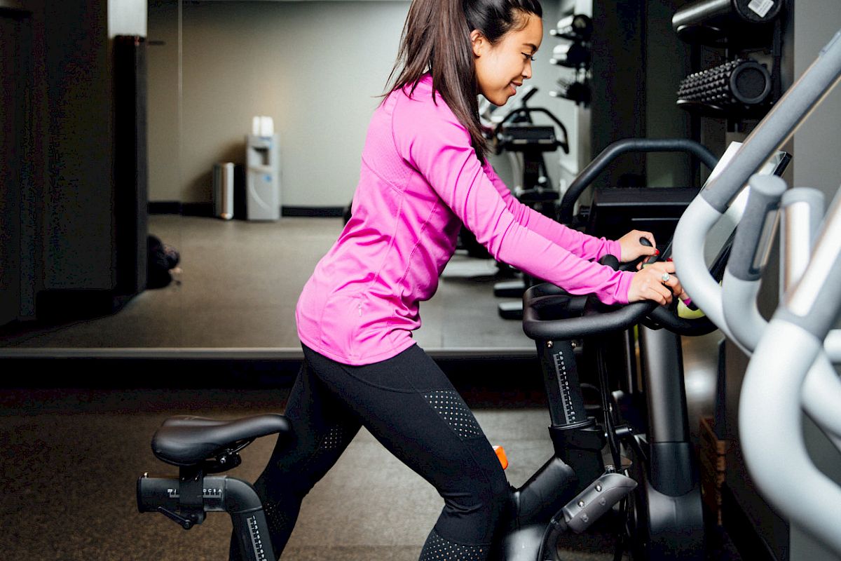 A woman in a pink top and black leggings exercises on a stationary bike in a gym. The background shows gym equipment and weights.
