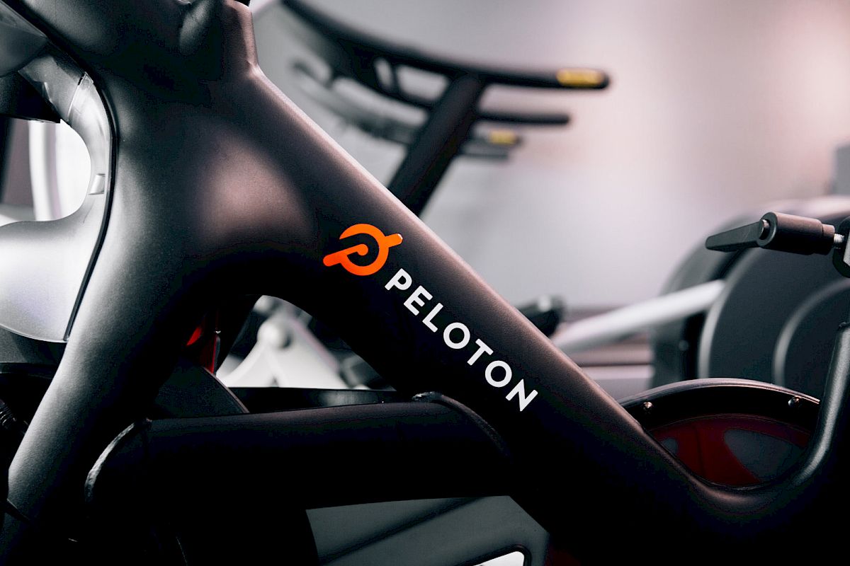 The image shows a close-up of a Peloton exercise bike, showcasing the brand name and logo on the frame in a gym setting.