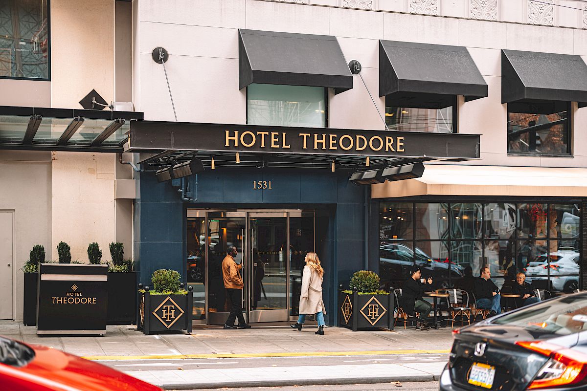 People walking by the entrance of Hotel Theodore, which features a modern exterior with a few plants, black awnings, and a busy street nearby.