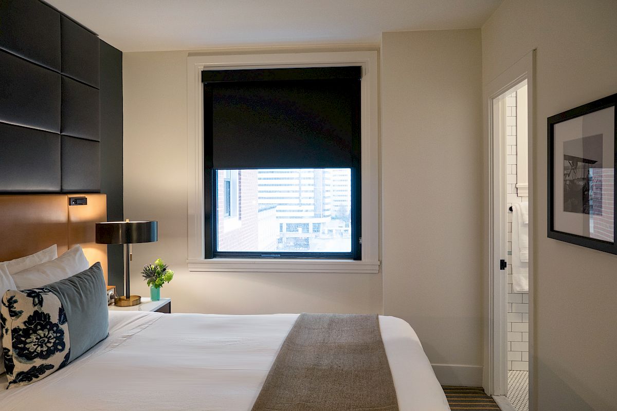 A neatly made bed with decorative pillows, a bedside table with a lamp and plant, a window with blinds, and a door leading to a tiled bathroom.