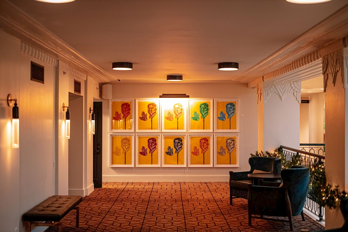 A hallway features a colorful artwork display with framed portraits, ambient lighting, a bench on one side, and chairs with a small table on another.