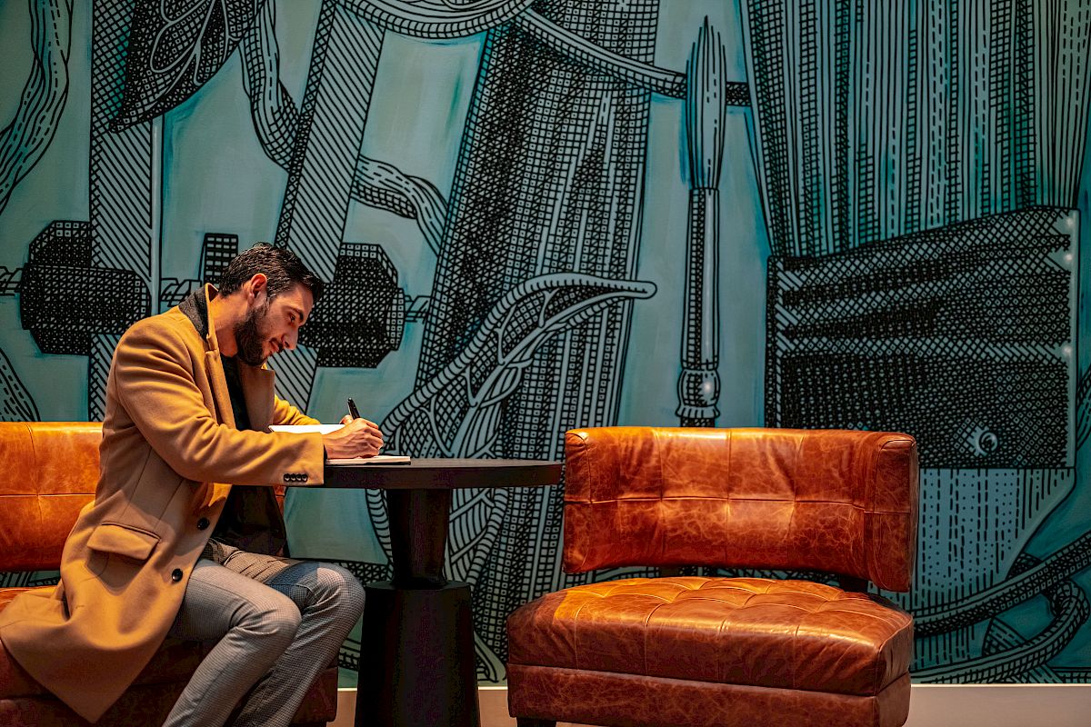 A man sits at a small table between two chairs, writing in a notebook, with an intricately decorated teal wall in the background.