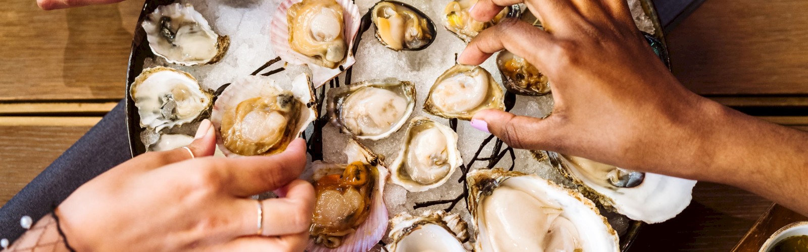 Three pairs of hands reach for a platter of oysters and grilled lemon halves, laid out on ice, with a variety of dipping sauces on the side.