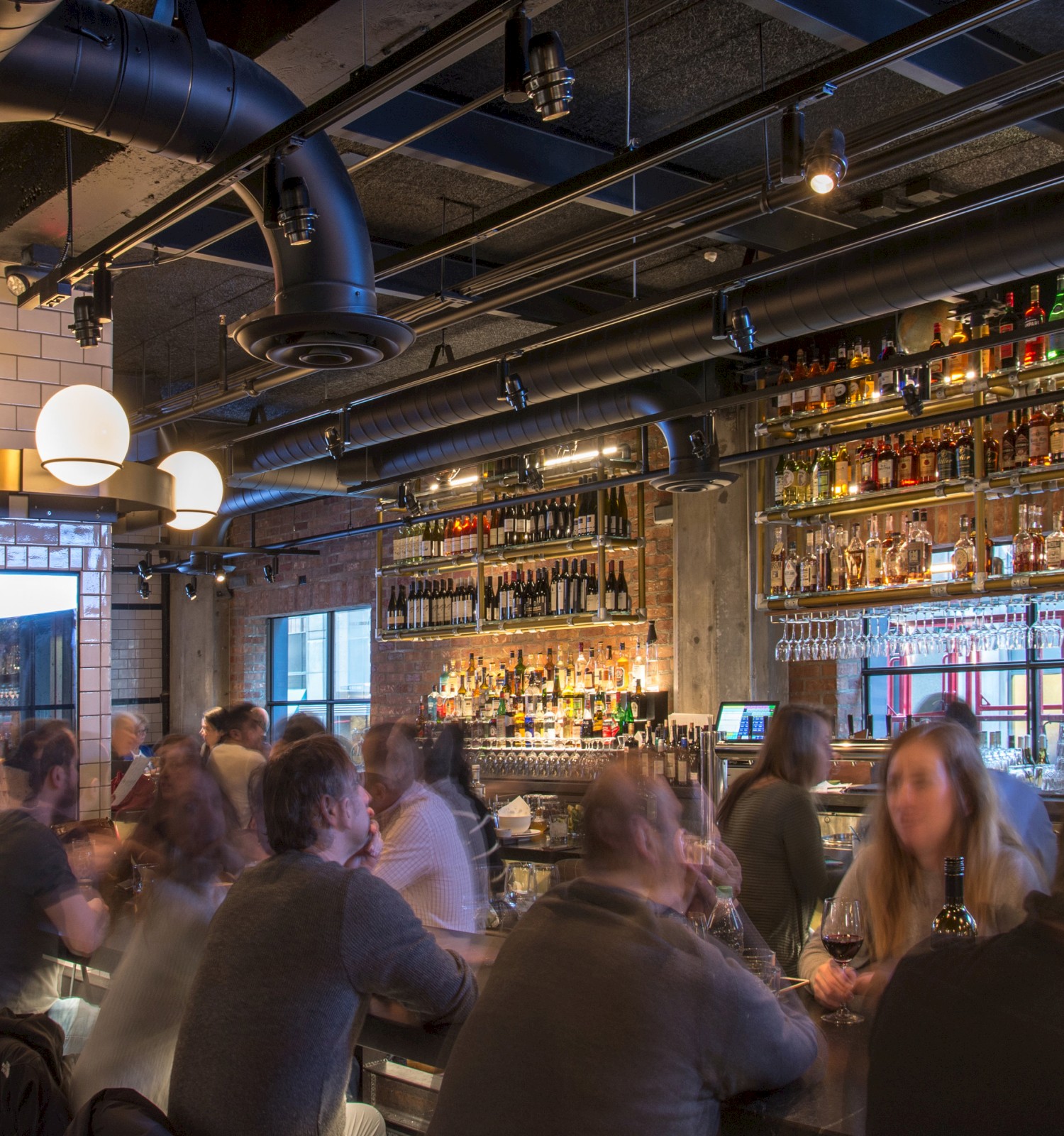 A busy bar with patrons sitting at the counter, bartenders working, and shelves stocked with various bottles of liquor behind the bar.
