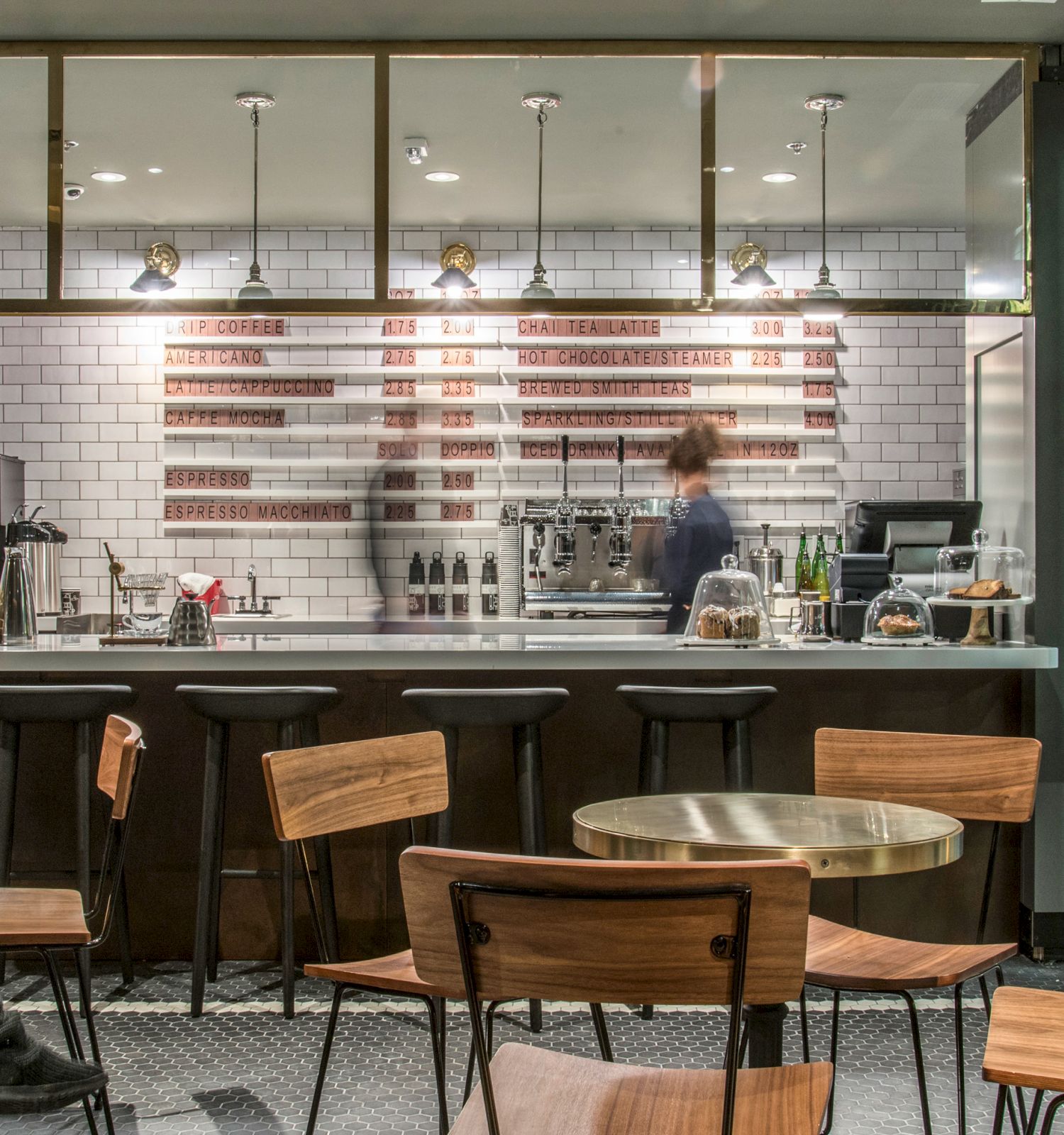 A cozy cafe with wooden chairs, a counter with bar stools, and a barista working behind it. A partial U.S. flag decorates the back wall.