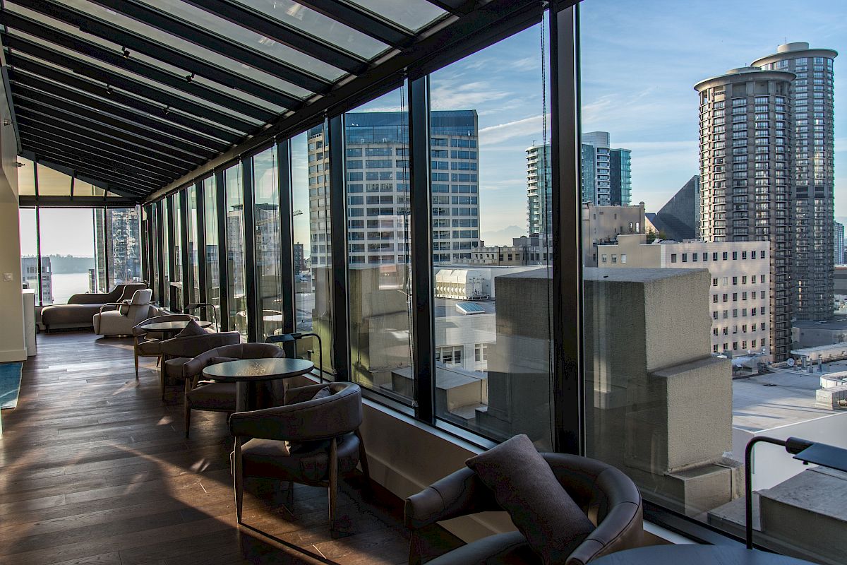 A modern lounge with glass walls offering a panoramic city view; it features comfortable seating and tables, creating a relaxing atmosphere.
