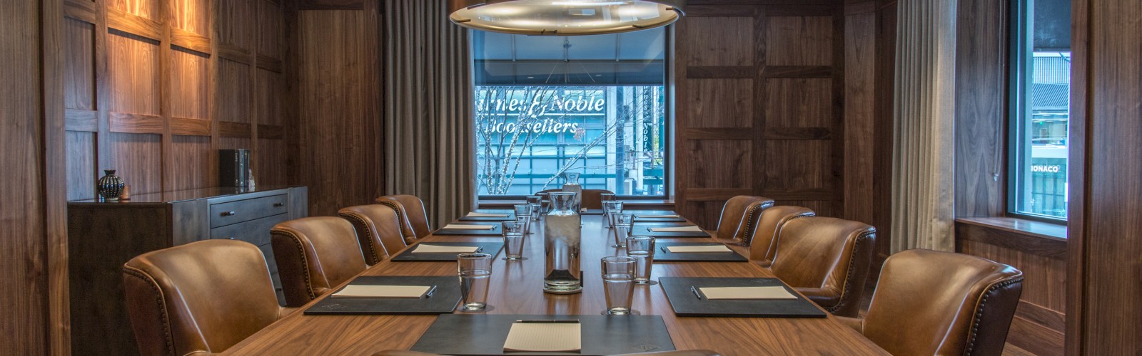 A well-furnished conference room with a large wooden table, leather chairs, notepads, and modern overhead lights, featuring wooden walls and large windows.