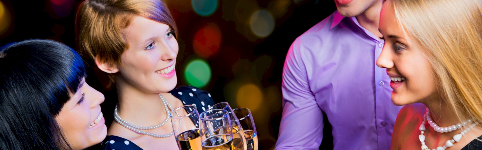 Four people are toasting with wine glasses at a party. Background features festive, colorful lights.