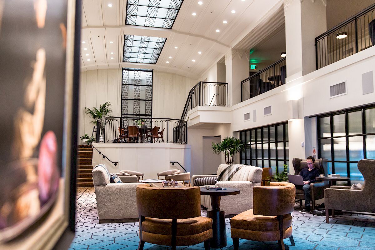 A spacious, modern lounge area with comfortable seating, high ceilings, and a mezzanine level, featuring a person sitting and working on a laptop.