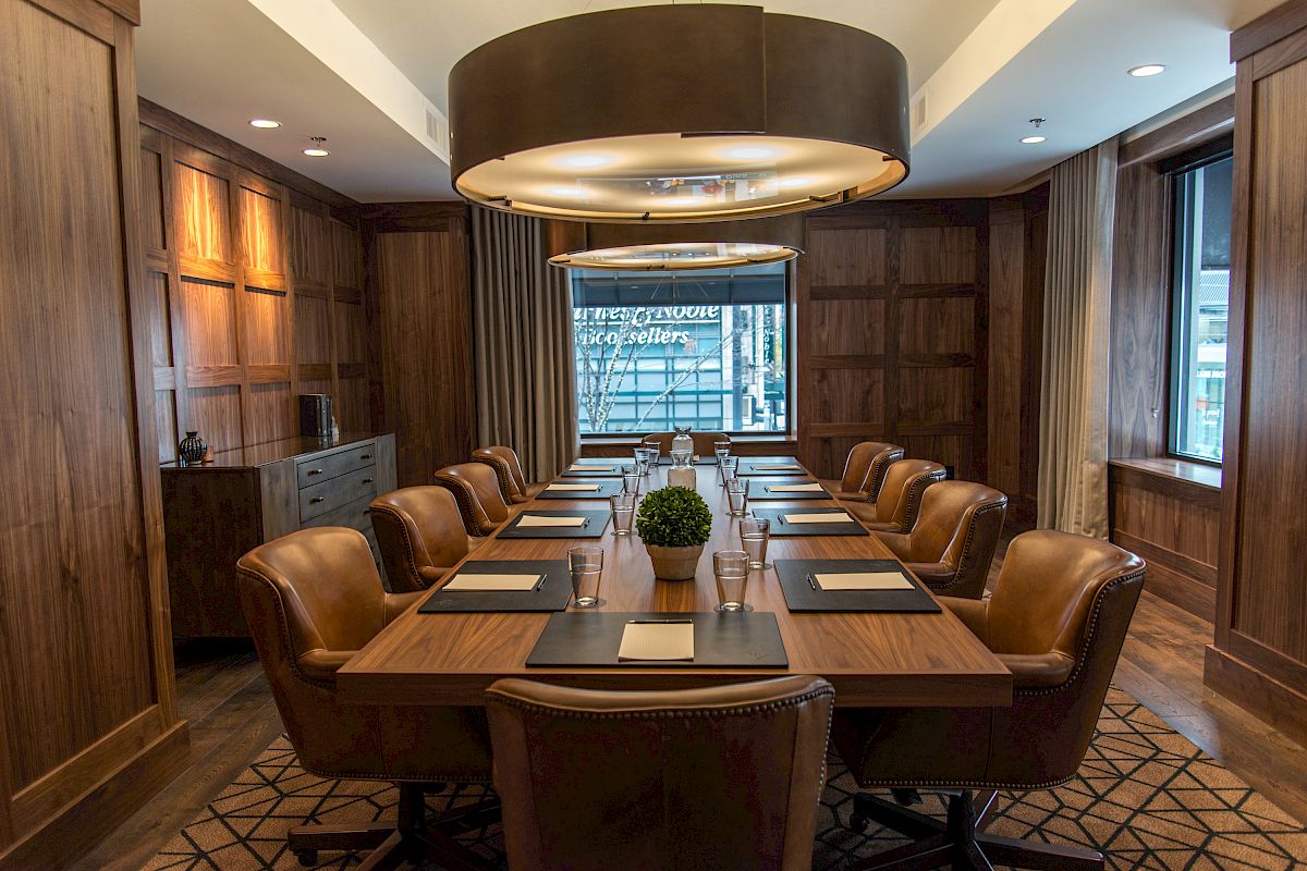 A modern conference room with a long wooden table, leather chairs, a central plant, and large, circular overhead lights. The walls are wood-paneled.