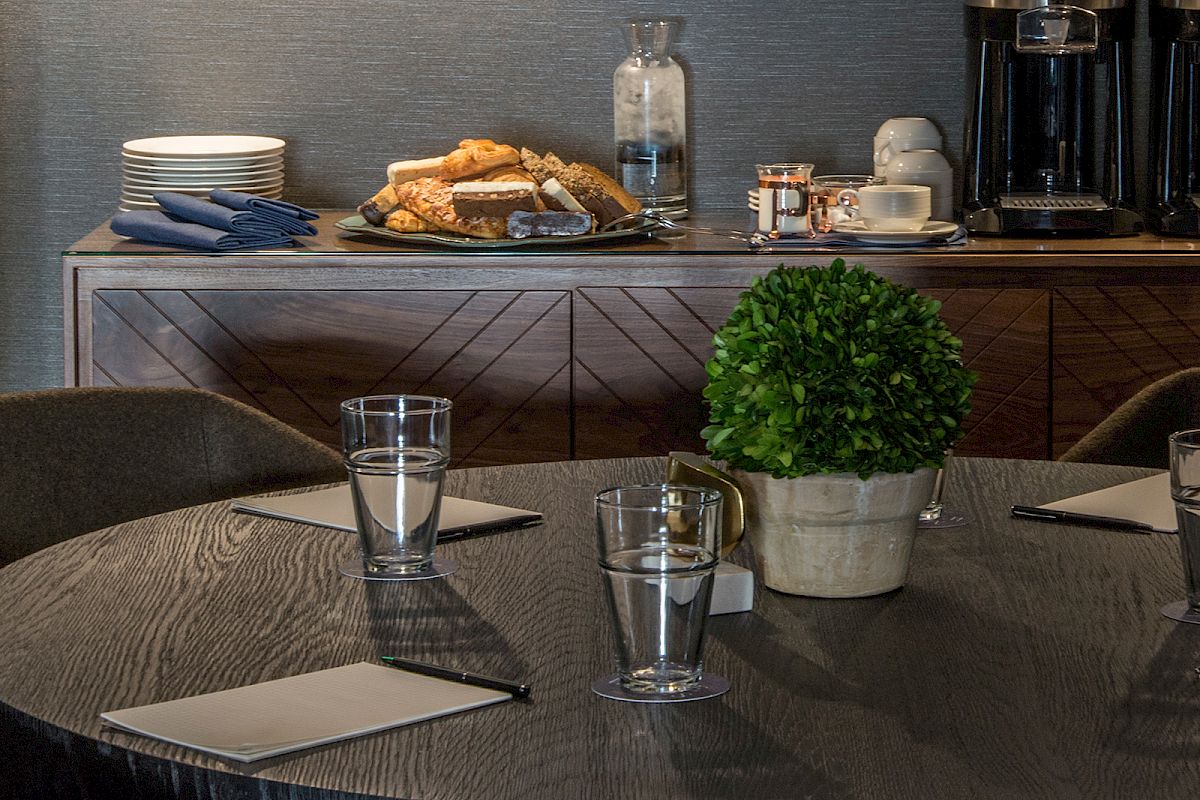 A table with glasses of water, notepads, a potted plant; in the background a sideboard with snacks, glasses, a coffee maker, and plates.