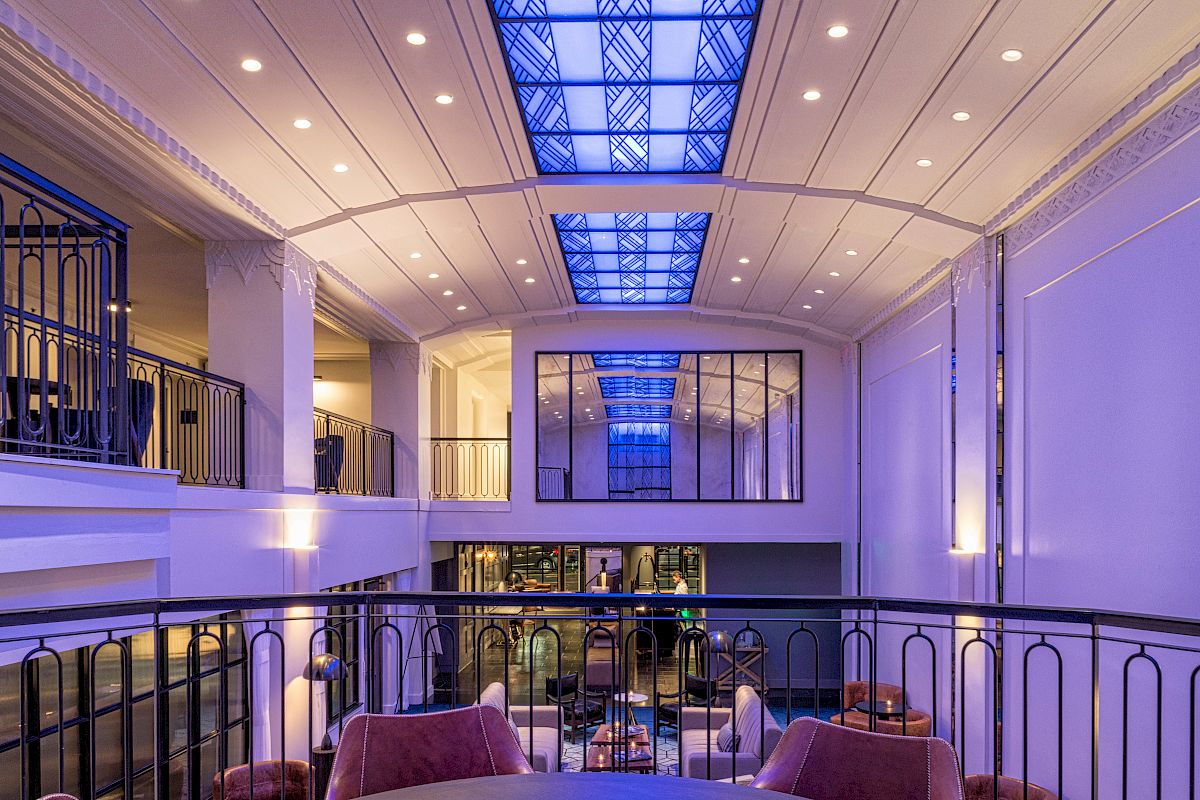 A modern, well-lit interior with a high ceiling, a glass skylight, and balcony seating overlooking a lounge area with chairs and tables.