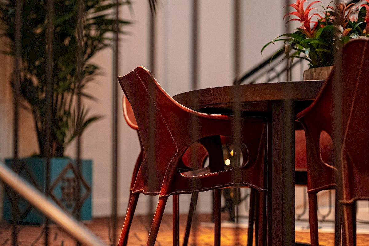 A cozy indoor space with a wooden table, red chairs, potted plants, and warm lighting, enclosed by a decorative railing.
