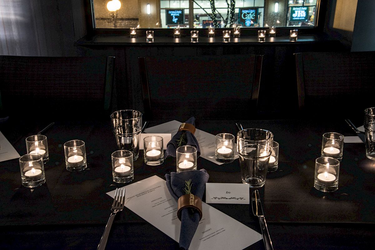 The image shows a dimly lit dining table set with candles, glasses, napkins, and menus, creating an intimate and cozy atmosphere.