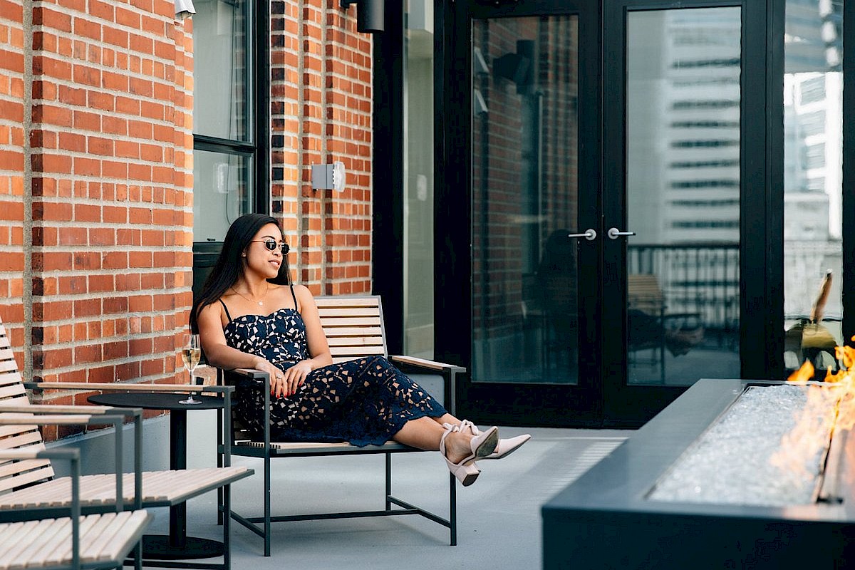 A woman sits on a patio near a brick wall and modern outdoor furniture, looking relaxed by a fire pit at a rooftop or balcony.